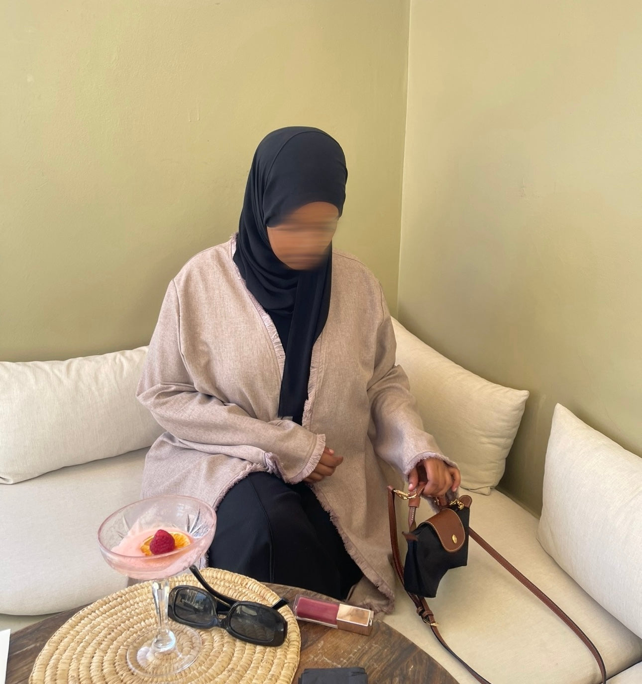 Person sitting on a couch with a beige linen open tassel abaya with a black hijab and a black mini longchamp bag and sunglasses