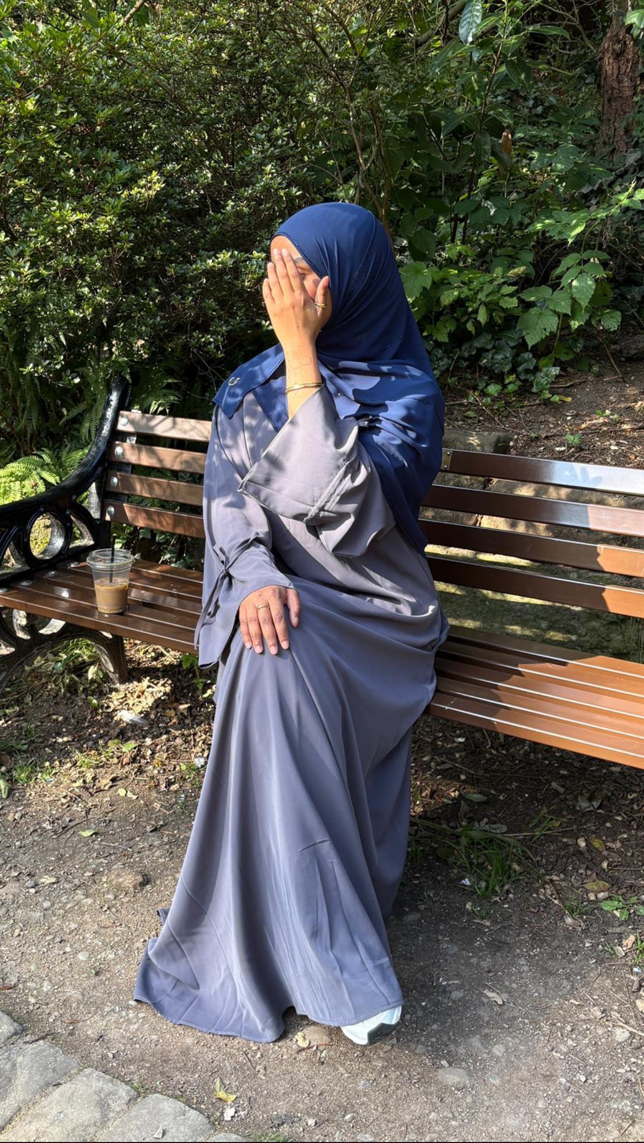 Woman in a blue hijab and grey nursing friendly abaya dress sitting on a bench in a park.