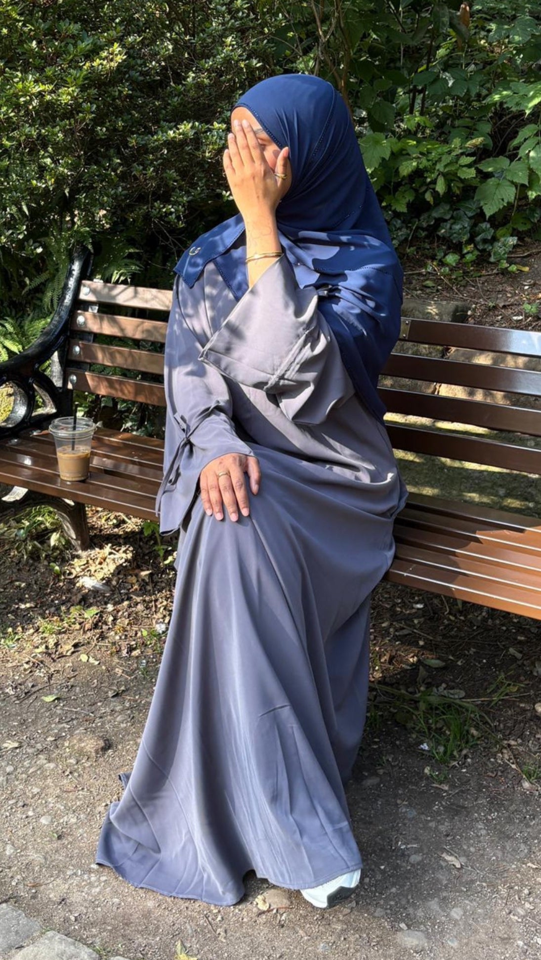 Woman in a blue hijab and grey nursing friendly abaya dress sitting on a bench in a park.