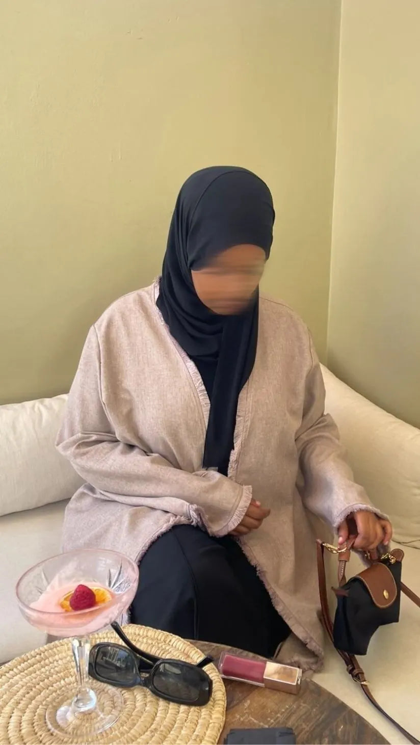 Person sitting on a couch with a drink and makeup items, wearing a hijab and beige linen tassel abaya